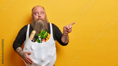 Chubby bearded adult man eats unhealthy food, points finger aside on copy space, tells where he bought all these products, leads junk lifestyle, has obesity problem, isolated on yellow background photo