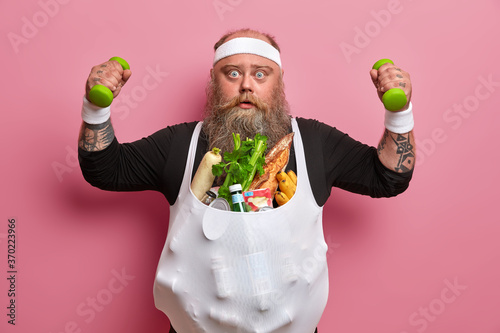 Surprised stout bearded man suffers from obesity caused by fast food addiction, burns calories and loses weight in sport gym, trains muscles with dumbbells, being motivated. Problem of gluttony photo