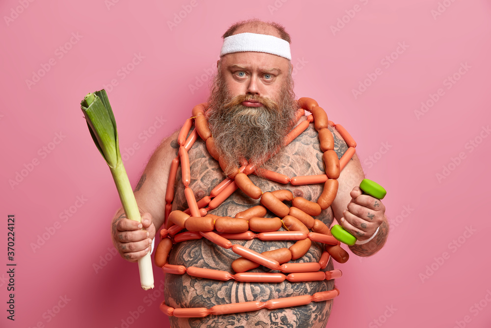 Unhappy obese fatso man wears white sports headband, holds dumbbell and green leek, has tattooed naked body wrapped in sausages, chooses between healthy and unhealthy food, has workout session