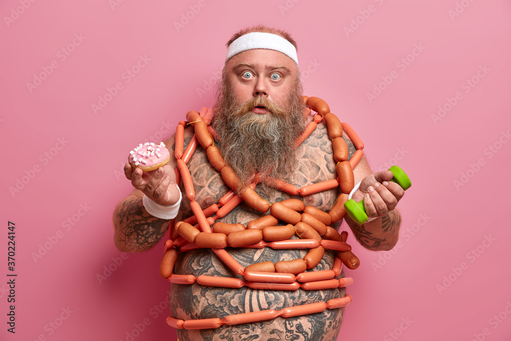 Stupefied stout bearded man poses with junk food and dumbbell, needs proper nutrition and sport exercises for losing weight, has fat stomach, holds donut, many sausages around naked tattooed body
