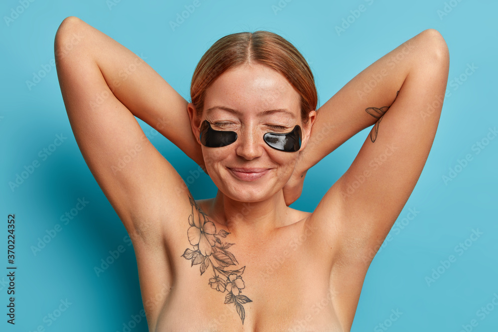 Beautiful sincere freckled woman keeps hands behind neck, closes eyes, applies black collagen patches, enjoys rejuvenation procedure, facial skin care product, stands topless with tattoo on body