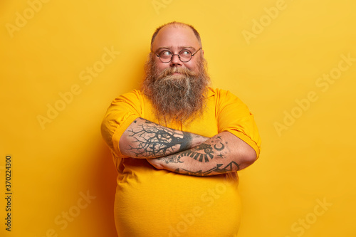 Φωτογραφία Handsome bearded man keeps arms folded, looks thoughtfully away, has plump body, dressed in casual clothes, makes up plan how to lose weight, isolated over yellow background