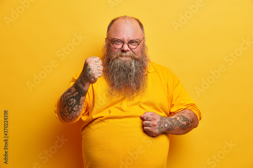 Photo of serious angry man has thick beard, clenches fists and looks with outraged expression, promises to revenge, shows stout big belly, dressed in yellow t shirt, expresses negative emotions