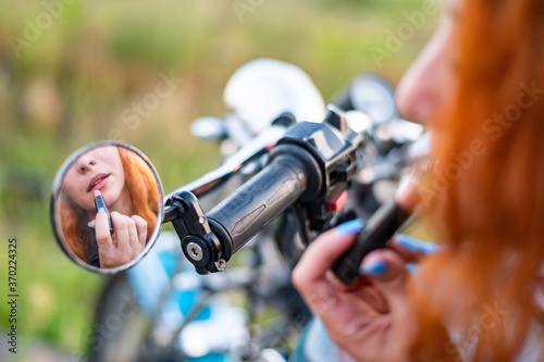 A woman on a motorcycle looks in the mirror and paints her lips with lipstick. Reflection of a red-haired biker girl.