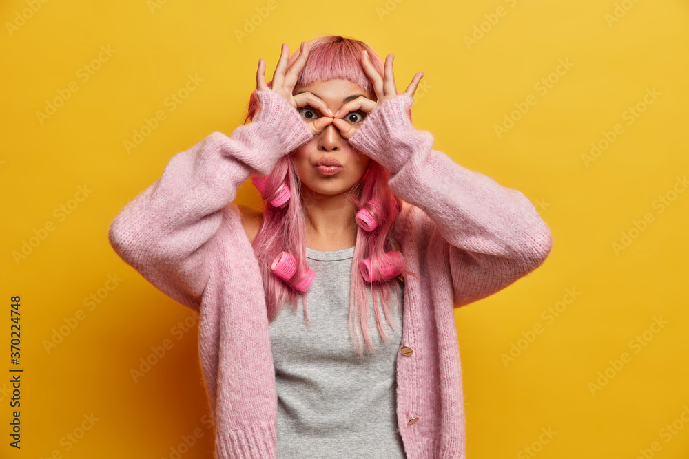 Studio shot of lovely surprised woman with pink hair, makes orbs with fingers, wears hair rollers for making perfect curls, peeks as wearing goggles, feels like child, enjoys life, shows okay gesture Studio shot of lovely surprised woman with pink hair, makes orbs with fingers, wears hair rollers for making perfect curls, peeks as wearing goggles, feels like child, enjoys life, shows okay gesture
