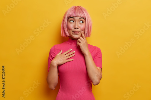 Photo of smiling happy Asian woman wears pink hair wig, imagines something pleasant, looks with dreamy peaceful expression, recalls good memories and makes wish, dressed in rosy casual clothes