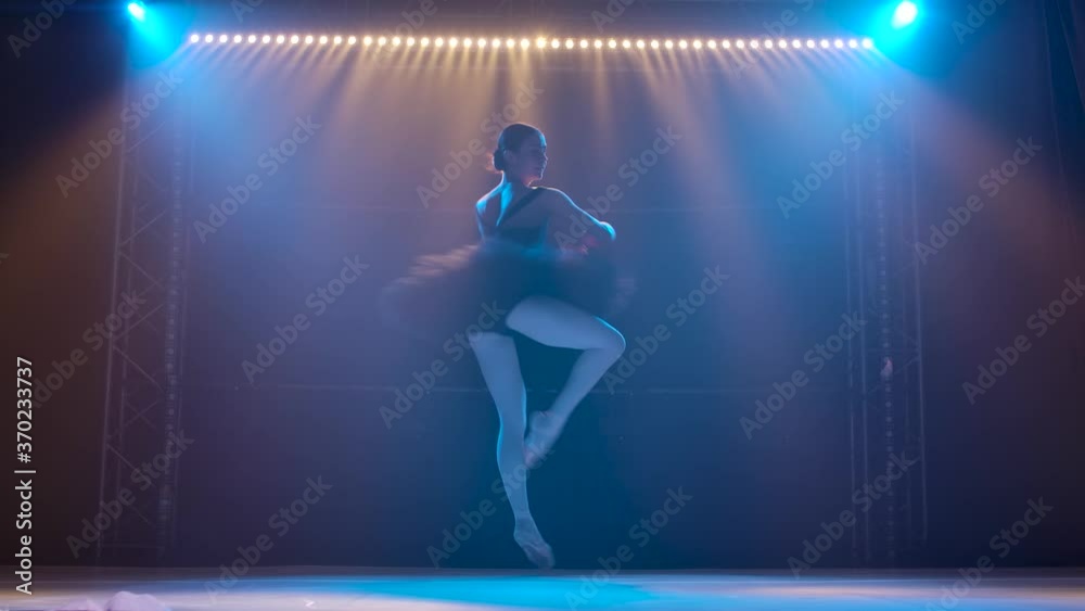 Silhouette a young flexible ballerina in black tutu and pointe ballet shoes. Graceful dancing of elements classical ballet. Shot in a darkness under a theatrical spotlights of studio. Slow motion.