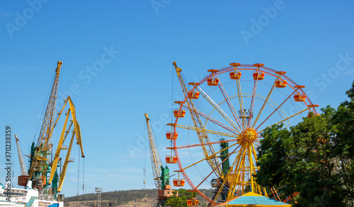 Summer day.A yellow Ferris wheel against a blue sky, next to portal cranes.