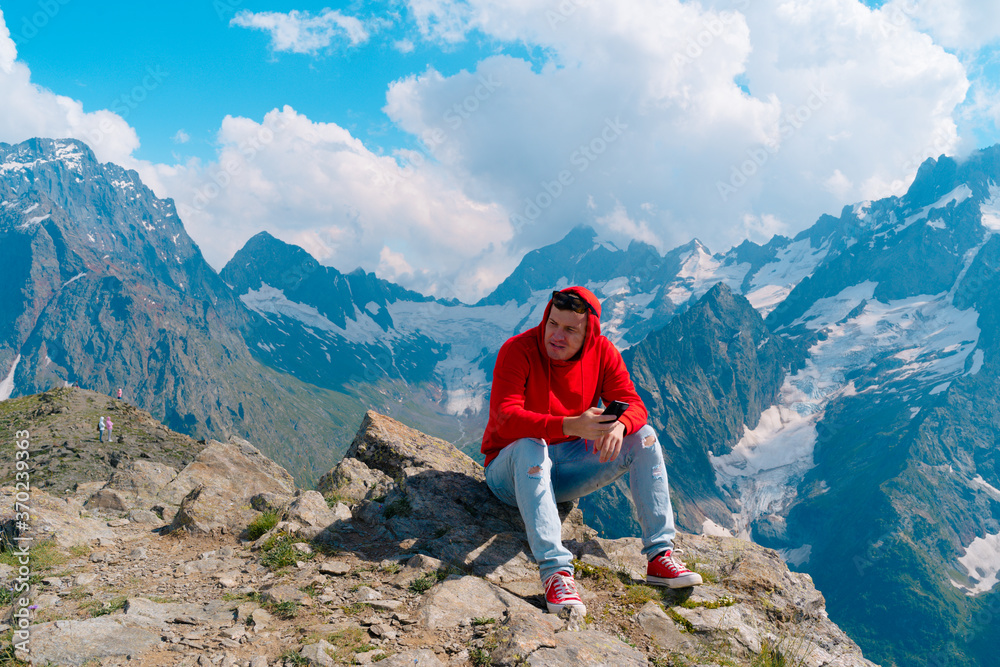 Man tourist sitting on rock with smartphone in mountains. Male traveler using mobile phone against cloudy sky on sunny day in mountainous terrain.