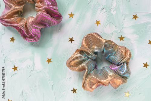 Two trendy holographic iridiscent shiny metallic scrunchies and golden stars confetti on green background. Diy accessories and hairstyles concept, copy space