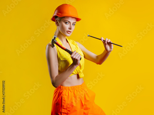 Fototapeta Young woman hammering nail at workshop on yellow background isolated