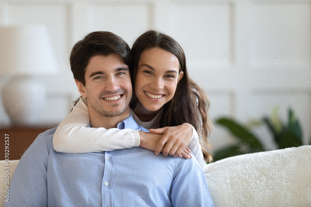 Portrait of happy beautiful young woman embracing smiling handsome man sitting on sofa. Bonding affectionate loving family couple homeowners looking at camera, relaxing together in living room. Portrait of happy beautiful young woman embracing smiling handsome man sitting on sofa. Bonding affectionate loving family couple homeowners looking at camera, relaxing together in living room.