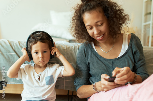 Indoor shot of attractive cheerful young Hispanic female holding mobile phone playing music tracks for her adorable baby son who listening to songs via wireless headphones. Technology and leisure