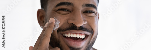 Close up head shot portrait overjoyed African American handsome young man applying moisturizing face cream, facial massage, enjoying skincare procedure, looking at camera, wide cropped image