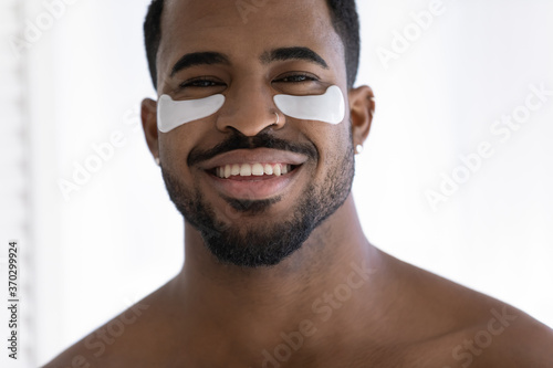 Head shot portrait close up smiling African American handsome young man with white hydrogel patches under eyes looking at camera, enjoying anti aging and dark circles skincare procedure