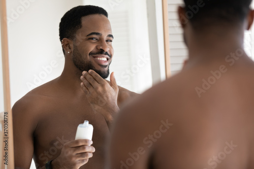 Mirror reflection close up smiling satisfied African American young man applying aftershave moisturizing lotion, standing in bathroom, enjoying skincare routine procedure after shaving photo