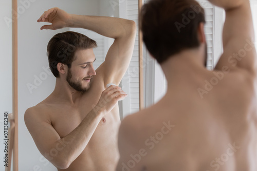 Mirror reflection young man applying antiperspirant on armpit after shower, standing in bathroom, handsome guy doing morning routine procedure, using stick underarm deodorant, personal hygiene