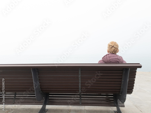 Fotografie Woman in despair sitting on a lonely bench and looking at grey distant horizon on a ocean foggy day
