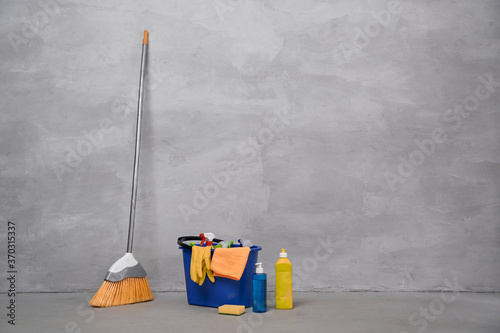 Cleaning equipment. Broom and plastic bucket or basket with cleaning products, bottles with detergents standing on the floor against grey wall