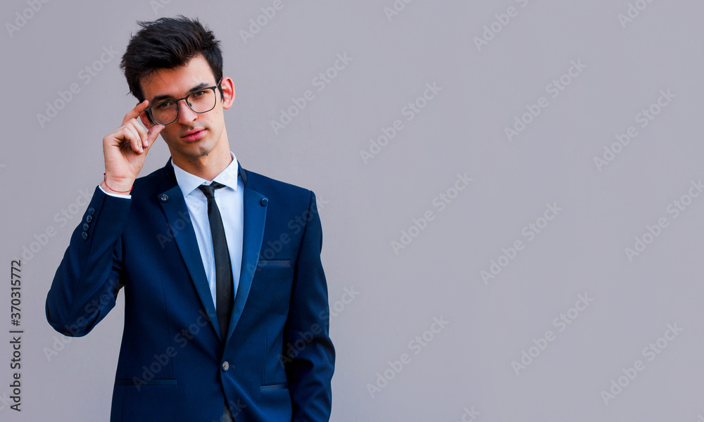 Young handsome man in blue suit and glasses smiling isolated on gray background Young handsome man in blue suit and glasses smiling isolated on gray background