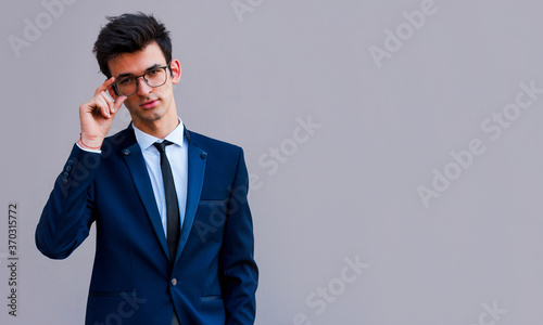Young handsome man in blue suit and glasses smiling isolated on gray background