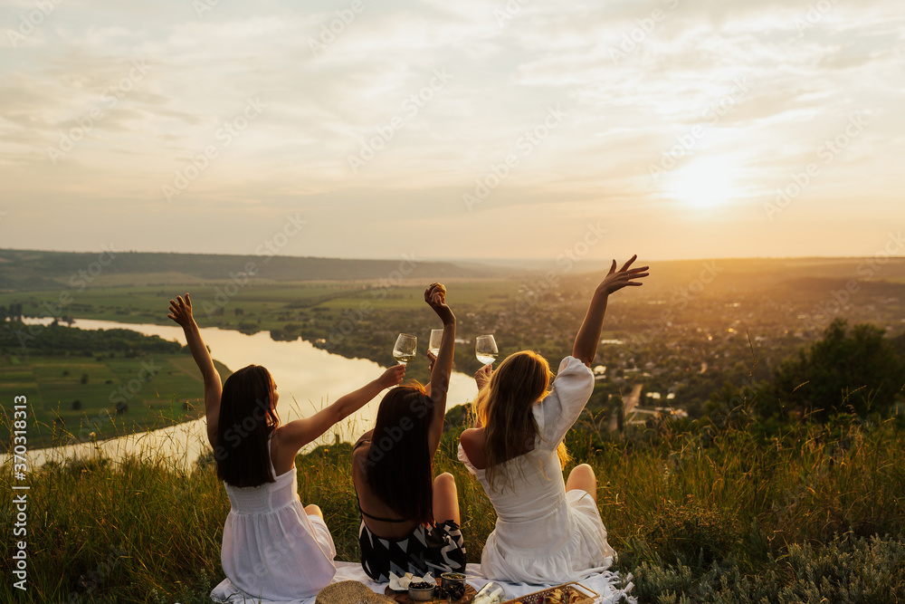 Three best girlfriends having fun together on a picnic on the mountain at sunset. They enjoying time in nature at weekend. Copy space.