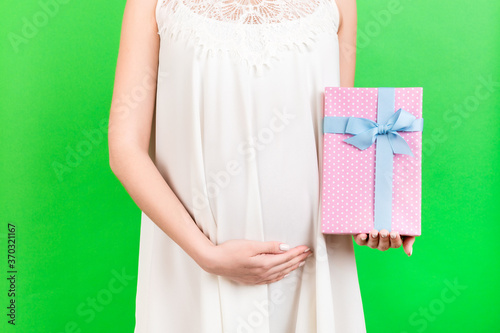 Close up of pregnant woman in white dress holding pink spotted gift box at green background. Expecting a baby girl. Copy space