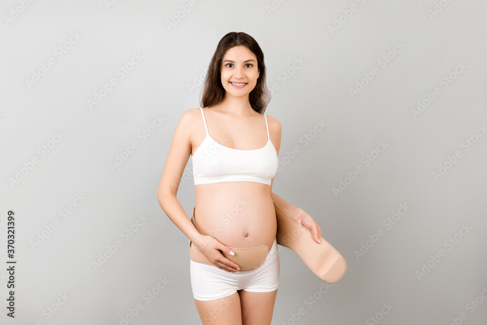 Portrait of pregnant woman in white underwear dressing maternity belt at gray background with copy space. Support belt against backache. Orthopedic abdominal support belt concept Portrait of pregnant woman in white underwear dressing maternity belt at gray background with copy space. Support belt against backache. Orthopedic abdominal support belt concept