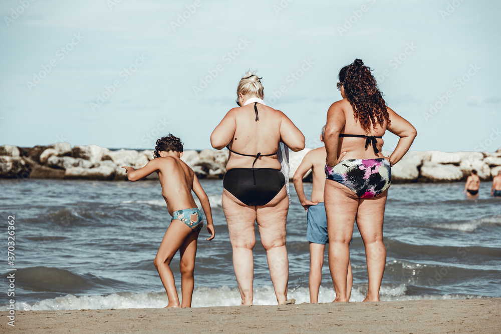 Overweight woman, mother and daughter, standing on the shore in a popular beach in Italy and looking at the sea with their arms on their hips. Vintage atmosphere.