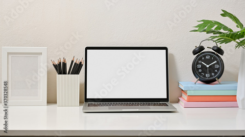 Mock up of creative workspace with laptop, pencils, frame, clock, and books on white table. Blank screen laptop.