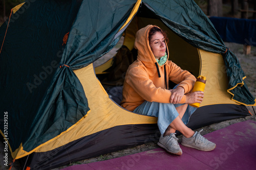A serene Caucasian woman sits alone in a tourist tent drinking hot tea from a thermos. The girl enjoys a drink from the thermo mug in the campground, watching the sunset.