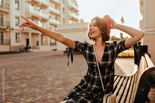 Slim romantic girl sitting on bench with smile. Outdoor shot of ecstatic french female model pointing finger at something.