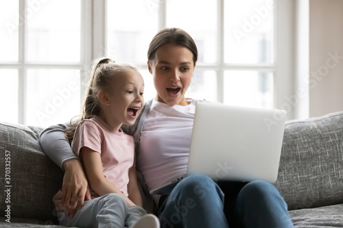 Happy young mother and little schoolgirl daughter looking at laptop screen, amazed by good discounts sale offers in internet store, involved in online shopping, surprised family watching funny movie.