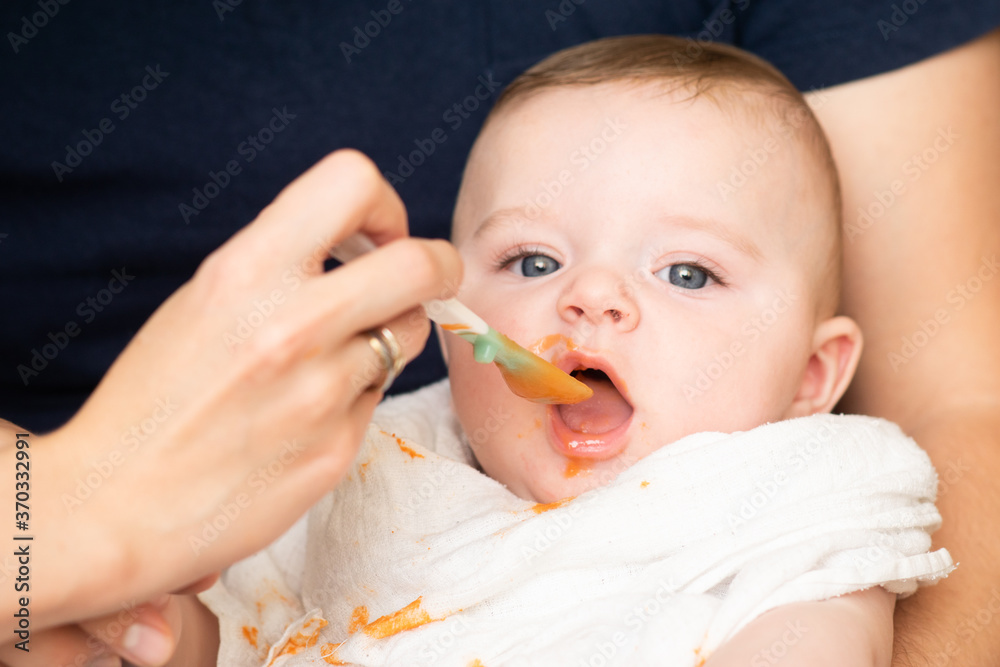 Tiny baby boy eating his vegetable carrot meal for the first time. Parents feeding a baby with a spoon - expanding diet. Tiny baby boy eating his vegetable carrot meal for the first time. Parents feeding a baby with a spoon - expanding diet.
