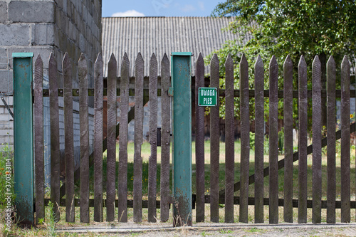 Drewniana furka z tabliczką informacyjną, "UWAGA PIES" oraz fragment bramy wjazdowej, wieś.
