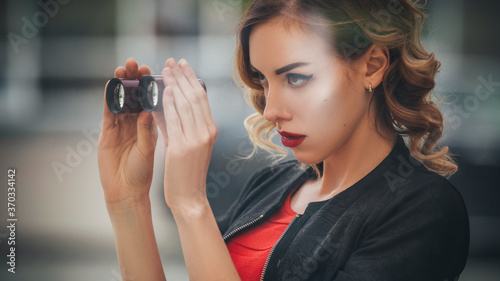 Beautiful brunette sexy spy agent (killer or police) woman in leather jacket and red luxury dress with a binocle in her hand watching after someone in european city photo