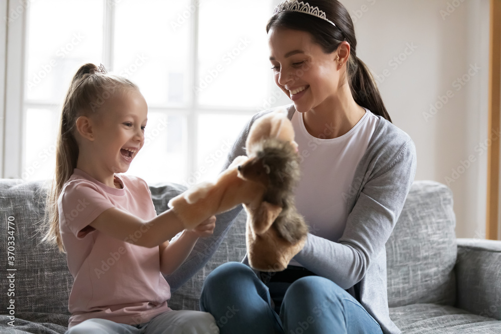 Overjoyed little adorable preschool blond girl playing hand toys with loving young mother, spending leisure time together indoors, relaxing on sofa. happy different generations family pastime concept.