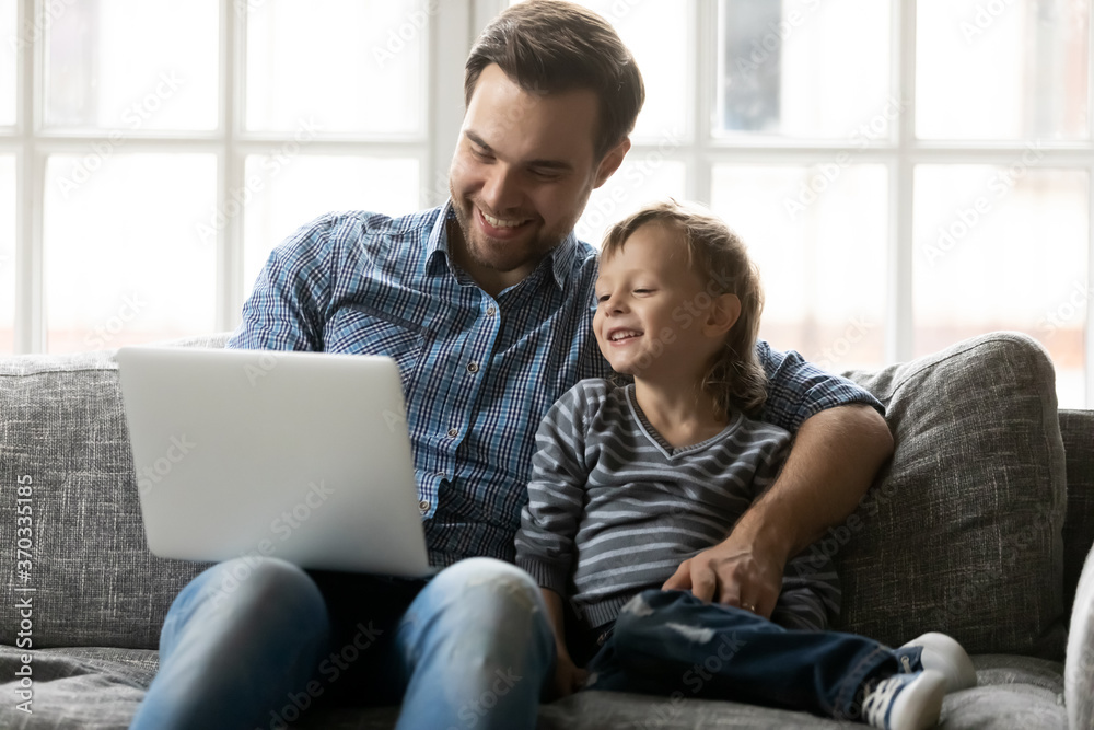 Smiling young handsome father embracing little preschool child son, using computer together in living room. Happy two generations family enjoying watching funny cartoons, movie or TV series at home.