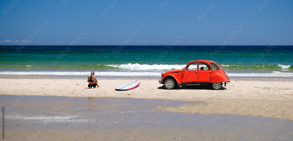Rapariga surfista sentada na praia com a prancha de surf pousada na areia e um carro antigo vermelho parado perto do mar, costa marítima, desporto radical, estilo de vida Rapariga surfista sentada na praia com a prancha de surf pousada na areia e um carro antigo vermelho parado perto do mar, costa marítima, desporto radical, estilo de vida