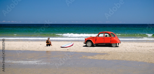 Rapariga surfista sentada na praia com a prancha de surf pousada na areia e um carro antigo vermelho parado perto do mar, costa marítima, desporto radical, estilo de vida