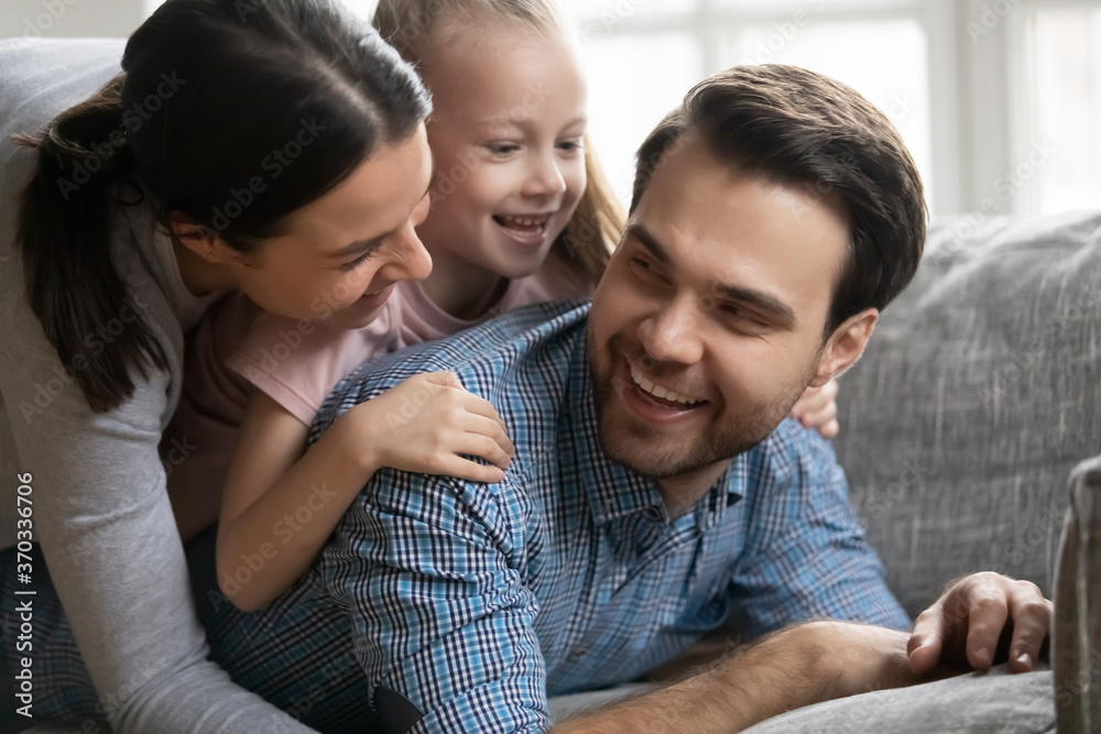 Happy young handsome man having fun with wife and adorable child girl, having fun together on comfortable sofa. Overjoyed hilarious couple enjoying spending free weekend time with small daughter.