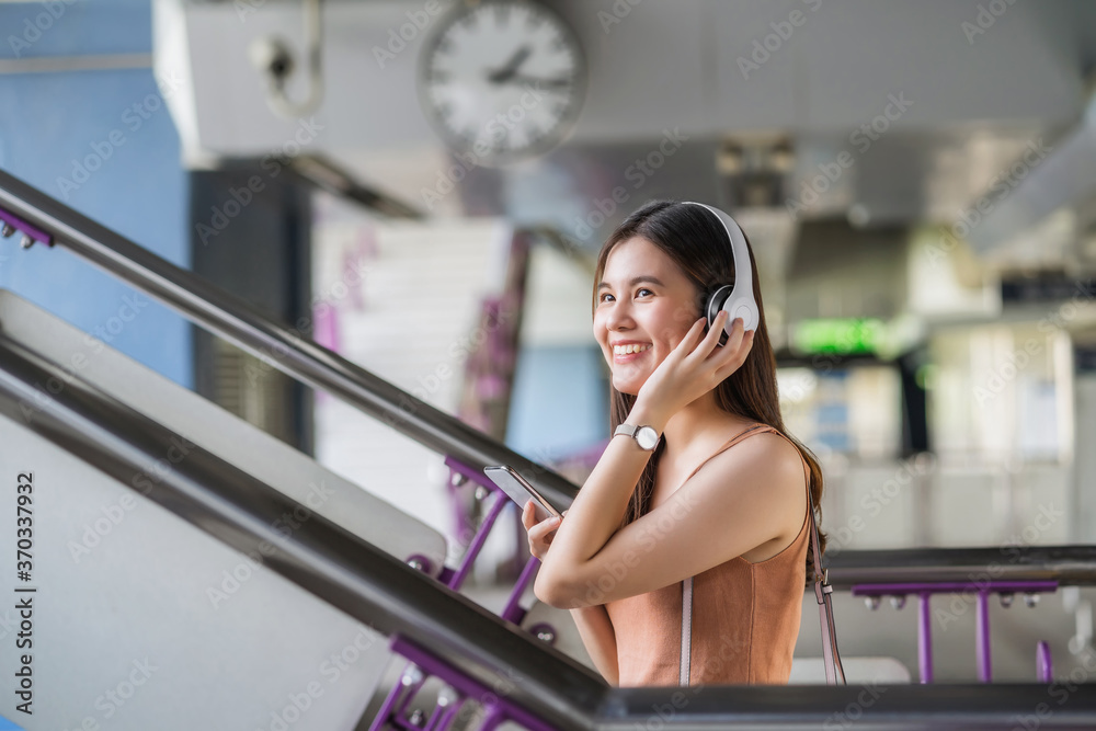 Young Asian woman passenger wearing headphone and using smart mobile phone to listening music and walking up the stairs in subway station when traveling in big city,commuter and transportation concept