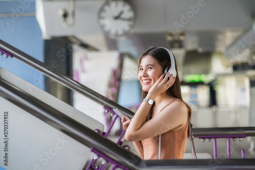 Obraz na płótnie Young Asian woman passenger wearing headphone and using smart mobile phone to listening music and walking up the stairs in subway station when traveling in big city,commuter and transportation concept w salonie