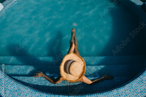 Top view over a young woman sitting in a pool, wearing a straw hat. Concept of summer holidays.