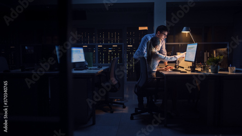 Ambitious Businessman Uses Desktop Computer, His Female Project Manager Explains Specific Tasks, Account Handling and Strategic Moves. Professional People Late at Night in Big Corporate Office