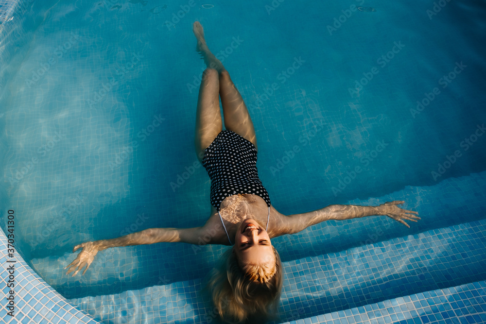 Young slim woman wearing a one-piece polka dot swimsuit, lying in a pool floating on water, outdoors.