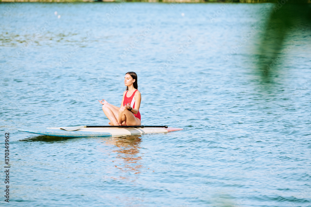 Young serene female in red swimsuit sitting on surfboard in pose of lotus