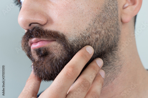 Foto Close up of young bearded man touching his beard while standing against gray bac