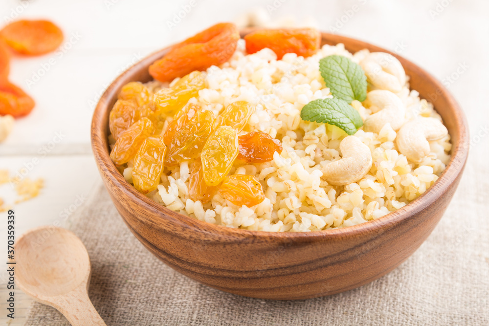 Bulgur porridge with dried apricots,  raisins and cashew in wooden bowl on a white wooden background and linen textile. Side view, selective focus.