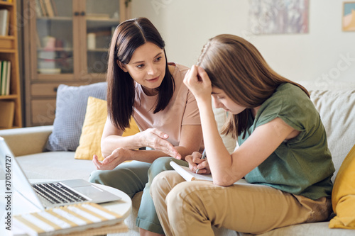 Young confident woman in casualwear helping her teenage daughter with homework